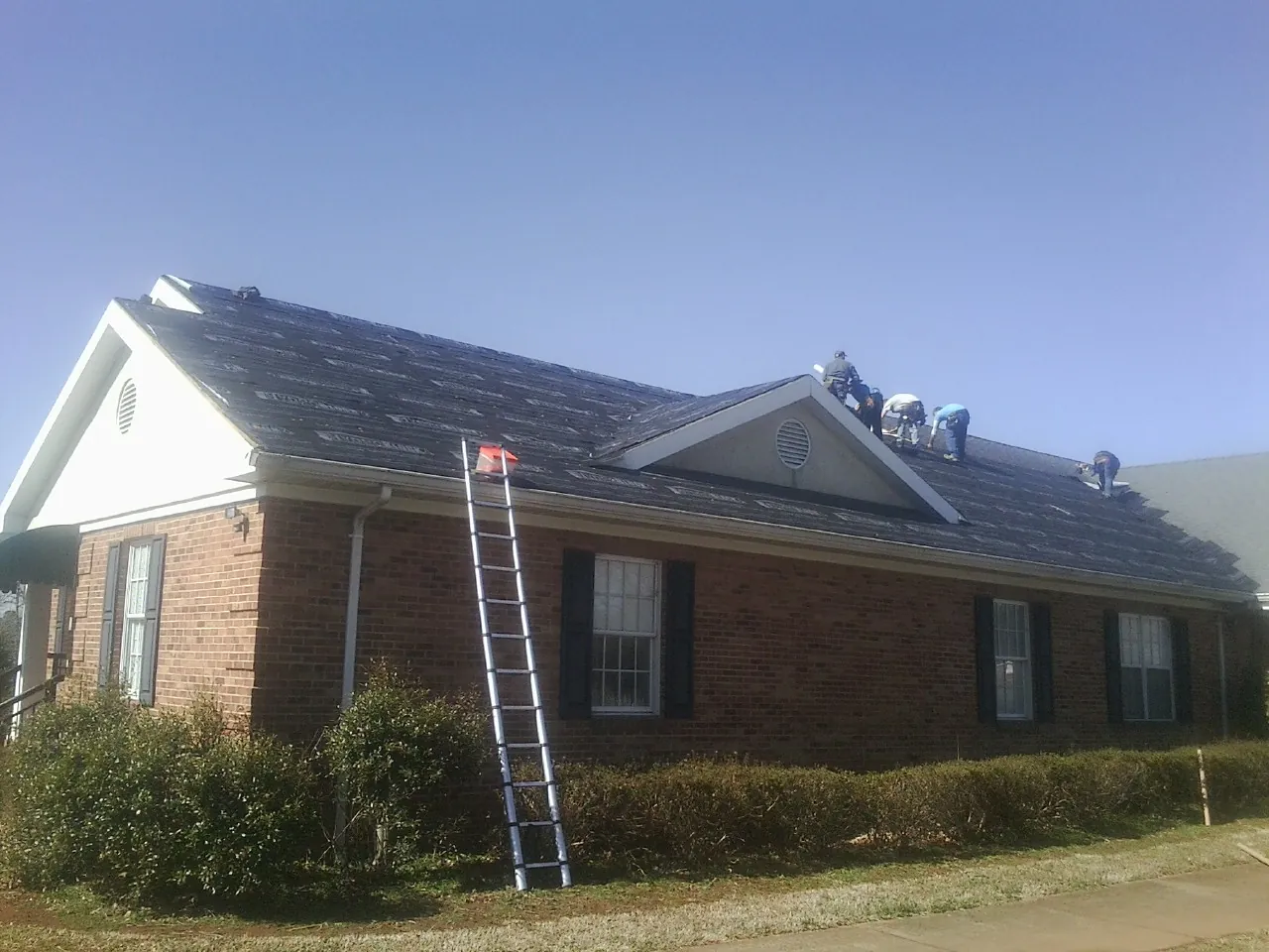 Crew tearing shingles off a brick ranch roof with a ladder set — Emergency Roof Repair work by our crew in Malone