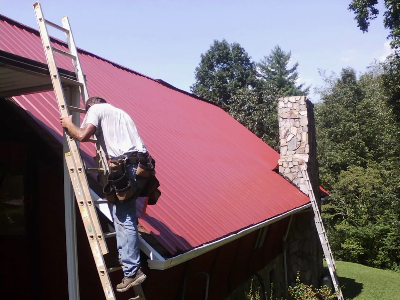Roofer on a ladder beside a red metal roof and stone chimney — Soffit & Fascia Repair by Roofing Brothers in Malone